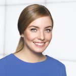 Eine Frau mit heller Haut, blauen Augen und glattem hellbraunem, nach hinten gezogenem Haar, die lächelt und ein blaues Oberteil trägt, posiert vor einem weißen Hintergrund mit einem dezenten Gittermuster.
