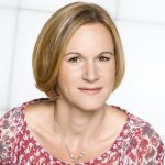 Eine Frau mit kurzen blonden Haaren lächelt sanft. Sie trägt ein rosa gemustertes Oberteil und eine silberne Halskette. Der Hintergrund ist hell und unscharf.