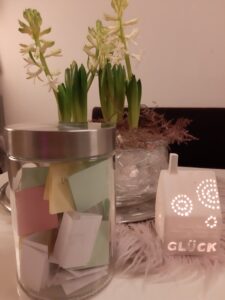 Ein Glasgefäß, gefüllt mit bunten gefalteten Papieren, steht auf einem Tisch neben frischen grünen und weißen Hyazinthenblüten in einer Vase und einem weißen Kerzenhalter mit ausgeschnittenem GLÜCK, der ein warmes Licht ausstrahlt.