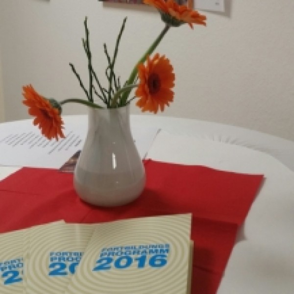 Eine kleine weiße Vase mit orangefarbenen Blumen steht auf einem Tisch mit roten Servietten und mehreren Broschüren mit der Aufschrift 2016. Im Hintergrund hängen bunte Bilder eines Kindergesichts an einer weißen Wand.