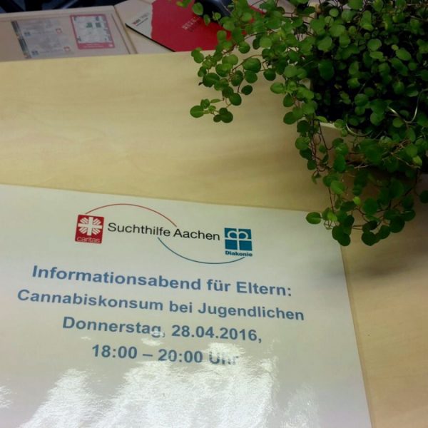 Ein Flyer auf einem Tisch neben einer Topfpflanze kündigt einen Informationsabend für Eltern zum Thema Cannabiskonsum unter Jugendlichen an, der am 28. April 2016 von 18:00 bis 20:00 Uhr in Aachen stattfindet.