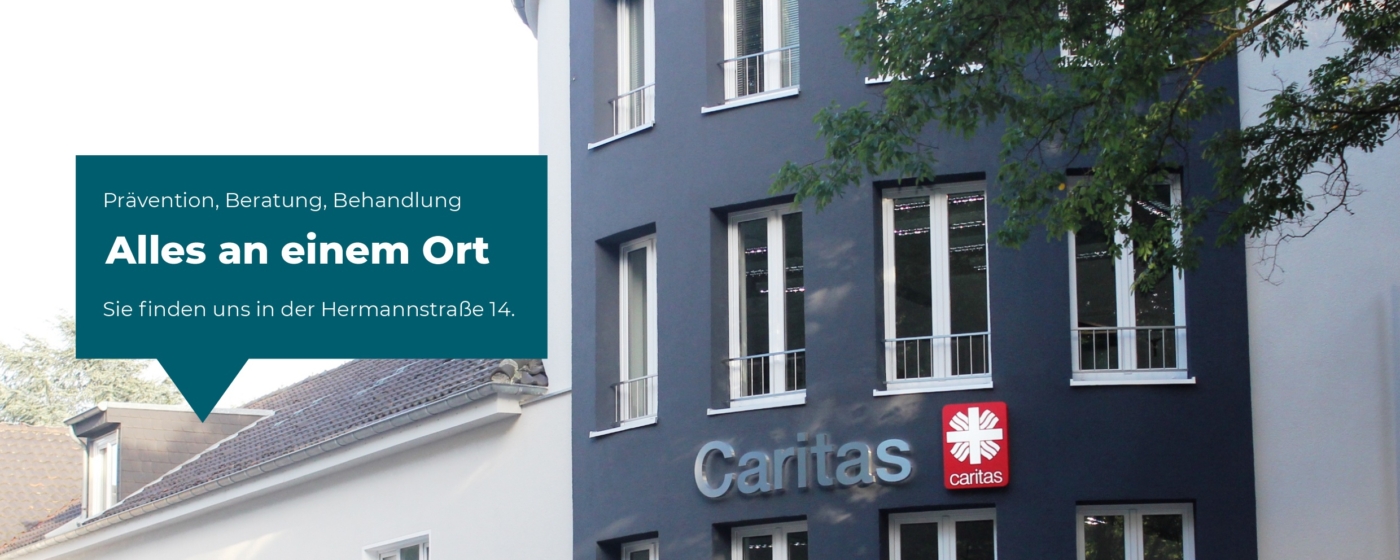 Auf einem Schild am Gebäude der Caritas steht: "Prävention, Beratung, Behandlung. Alles an einem Ort. Sie finden uns in der Hermannstraße 14." Das Gebäude hat große Fenster und das Caritas-Logo über dem Eingang.