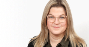 Eine Frau mit langen blonden Haaren und Brille lächelt in die Kamera. Sie trägt ein dunkles Oberteil und eine schwarze Jacke. Der Hintergrund ist einfarbig weiß.