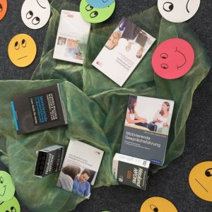 Mehrere Bücher über Kommunikationsfähigkeiten sind auf einem grünen Tuch angeordnet, umgeben von bunten Papierkreisen mit verschiedenen handgezeichneten Gesichtsausdrücken, wie lächelnde, neutrale und traurige Gesichter, auf einer dunklen Fläche.