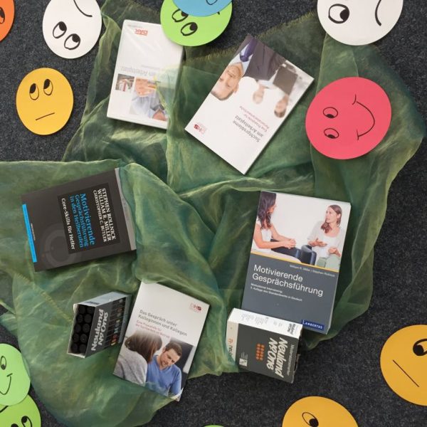 Mehrere Bücher über Kommunikationsfähigkeiten sind auf einem grünen Tuch angeordnet, umgeben von bunten Papierkreisen mit verschiedenen handgezeichneten Gesichtsausdrücken, wie lächelnde, neutrale und traurige Gesichter, auf einer dunklen Fläche.