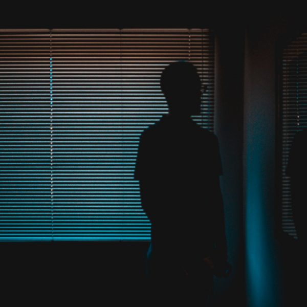 Die Silhouette einer Person, die in einem Innenraum vor einem geschlossenen Fenster steht, durch das gedämpftes blaues Licht dringt, schafft eine stimmungsvolle und geheimnisvolle Atmosphäre.