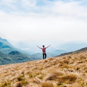 Eine Person steht mit erhobenen Armen auf einem grasbewachsenen Hügel und blickt auf eine weite Landschaft mit grünen Bergen und Tälern unter einem teilweise bewölkten Himmel.