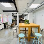Ein moderner, offener Büroraum mit einem hohen Holztisch, türkisfarbenen Hockern, Pflanzen auf dem Tisch, gefliesten Böden, weißen Wänden, großen Fenstern und sichtbaren Decken mit Industriebeleuchtung.