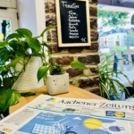 Ein Café-Interieur mit einer Topfpflanze auf einem Tisch, einer deutschen Zeitung im Vordergrund und einer Kreidekarte an einer Backsteinwand, auf der verschiedene Toastie-Füllungen zum Preis von 1 € aufgeführt sind.