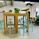 Helles, modernes Café mit hellen Fliesenböden, hohen Holztischen und türkisfarbenen Barhockern. Große Fenster lassen natürliches Licht herein. Topfpflanzen und verschiedene Stühle schaffen eine einladende, gemütliche Atmosphäre.
