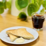 Ein weißer Teller mit zwei dreieckigen Scheiben Toast und einem Strudel aus Butter, neben einem Glas schwarzen Kaffee auf einem Holztisch. Grüne Blattpflanze verschwommen im Hintergrund.