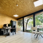 Modernes Büro mit Holzwänden, großen Fenstern, zwei Schreibtischen mit Computern auf der linken Seite und einem runden Tisch mit vier Stühlen auf der rechten Seite. Draußen sind grüne Bäume und Gebäude zu sehen. Natürliche und künstliche Beleuchtung.