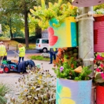 Bunt bemalte, mit Pflanzen gefüllte Blechdosen sind an einem Pfahl im Vordergrund befestigt; im Hintergrund arbeiten Menschen in gelben Westen in der Nähe eines Lieferwagens und eines kleinen Wagens in einer parkähnlichen Umgebung.