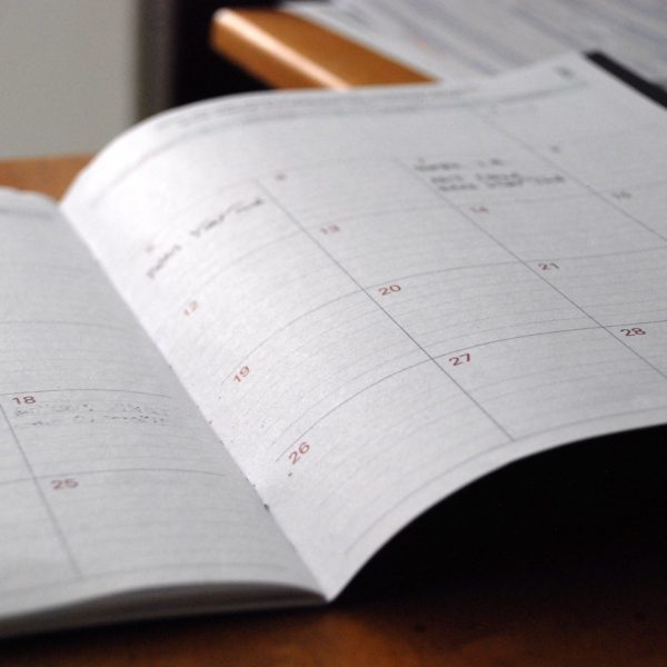 Ein offener Planer oder Kalender mit einem monatlichen Layout und leeren Datumsfeldern, der auf einem Schreibtisch mit einigen Papieren und einer sichtbaren Holzkante im Hintergrund liegt.
