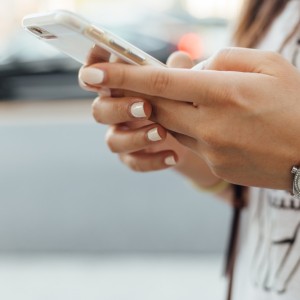 Eine Person mit ordentlich manikürten Nägeln hält ein Smartphone in der Hand und tippt. Sie trägt eine silberne Armbanduhr und ein weißes Hemd mit der Umrisszeichnung eines Elefanten. Der Hintergrund ist unscharf.