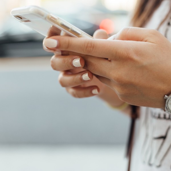Eine Person mit ordentlich manikürten Nägeln hält ein Smartphone in der Hand und tippt. Sie trägt eine silberne Armbanduhr und ein weißes Hemd mit der Umrisszeichnung eines Elefanten. Der Hintergrund ist unscharf.