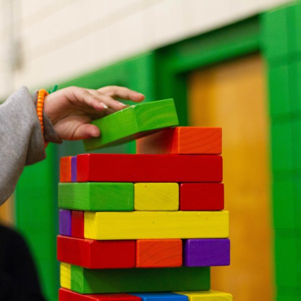 Die Hand einer Person legt einen grünen rechteckigen Block auf einen bunten Stapel von Holzblöcken, wahrscheinlich bei einem Stapelspiel, mit einer grünen und gelben Wand im Hintergrund.