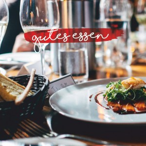 Nahaufnahme eines Gourmetgerichts auf einem weißen Teller, garniert mit Grünzeug, auf einem mit Brot, Weingläsern und Kerzen gedeckten Restauranttisch. Die Worte gutes essen erscheinen in handschriftlicher Schrift.