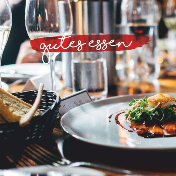Nahaufnahme eines Gourmetgerichts auf einem weißen Teller, garniert mit Grünzeug, auf einem mit Brot, Weingläsern und Kerzen gedeckten Restauranttisch. Die Worte gutes essen erscheinen in handschriftlicher Schrift.