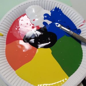 Ein weißer Pappteller mit roten, weißen, blauen, grünen, gelben, orangefarbenen und schwarzen Farbklecksen, die in Abschnitten angeordnet sind. Ein Pinsel mit blauer Farbe liegt quer auf dem Teller.