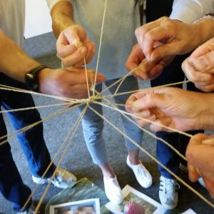 Mehrere Personen stehen in einem Kreis und halten sich an Schnüren fest, die in der Mitte zusammengebunden sind und Teamwork und Verbindung symbolisieren. Nur Hände und Arme sind sichtbar. Einige Fotos und Papiere liegen auf dem Boden darunter.