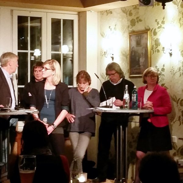 Sieben Erwachsene stehen und sitzen um Stehtische in einem warm beleuchteten Raum und unterhalten sich. Einige halten Mikrofone und Papiere in der Hand. Es scheint sich um eine informelle Diskussion oder eine Podiumsdiskussion in einem gemütlichen, dekorierten Raum zu handeln.