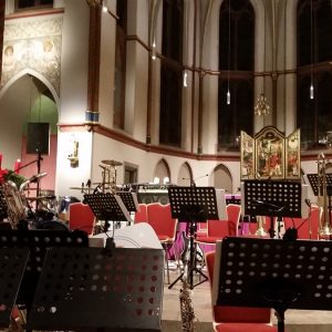 Ein leeres Orchester in einer Kirche mit roten Stühlen, Notenständern und verschiedenen Musikinstrumenten, darunter Schlagzeug und Blechbläser. Im Hintergrund sind Glasfenster, Kerzen und kunstvolle Dekorationen zu sehen.