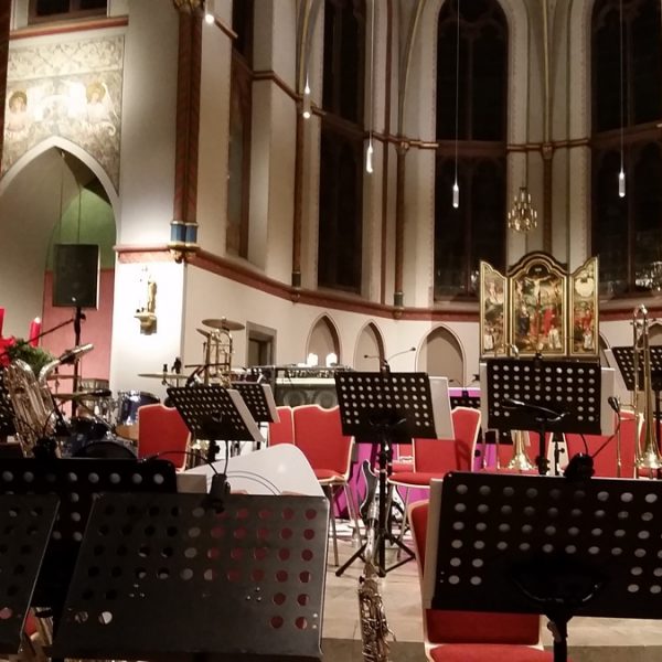 Ein leeres Orchester in einer Kirche mit roten Stühlen, Notenständern und verschiedenen Musikinstrumenten, darunter Schlagzeug und Blechbläser. Im Hintergrund sind Glasfenster, Kerzen und kunstvolle Dekorationen zu sehen.