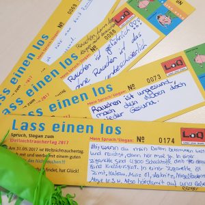 Vier gelbe und orangefarbene Lesezeichen mit der Aufschrift "Lass einen los" und Anti-Raucher-Botschaften in deutscher Sprache liegen auf einem Tisch. Ein grüner, nicht aufgeblasener Luftballon ist an einem Lesezeichen befestigt.