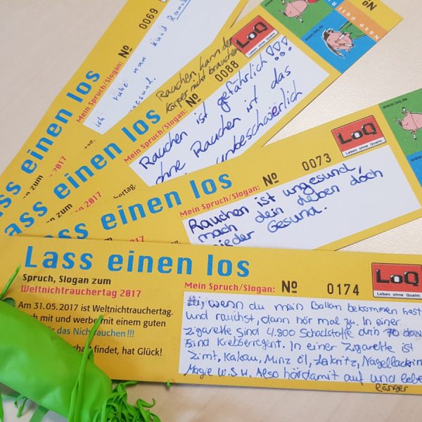 Vier gelbe und orangefarbene Lesezeichen mit der Aufschrift "Lass einen los" und Anti-Raucher-Botschaften in deutscher Sprache liegen auf einem Tisch. Ein grüner, nicht aufgeblasener Luftballon ist an einem Lesezeichen befestigt.