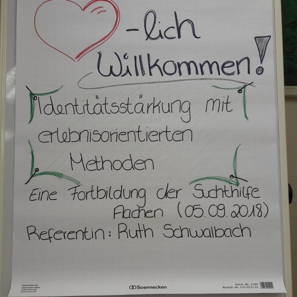 Ein Flipchart mit einem handgezeichneten roten Herz und dem Schriftzug lich Willkommen!, gefolgt von Details zu einem Workshop zur Identitätsstärkung in der Suchthilfe mit erlebnispädagogischen Methoden, geleitet von Ruth Schwalbach.