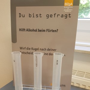 Drei durchsichtige Röhrchen stehen auf einem Tisch vor einem Schild in deutscher Sprache, auf dem gefragt wird, ob Alkohol beim Flirten hilft. Jedes Röhrchen ist mit "JA", "VIELLEICHT" oder "NEIN" beschriftet und enthält farbige Kugeln zur Abstimmung.