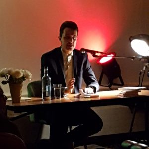 Ein Mann im Anzug sitzt an einem Schreibtisch mit einem Mikrofon, Papieren, einer Flasche Wasser und einer Lampe. Der Hintergrund ist schwach mit rotem Licht beleuchtet, was eine konzentrierte und intime Atmosphäre schafft.
