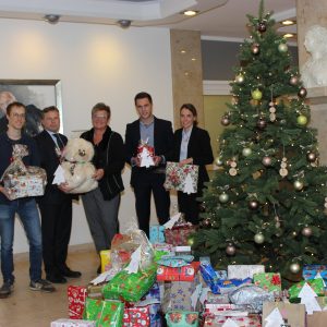 Fünf Personen stehen neben einem geschmückten Weihnachtsbaum, umgeben von vielen eingepackten Geschenken. Sie sind drinnen, halten Geschenke und lächeln in die Kamera. Im Hintergrund sind ein gerahmtes Porträt und eine Büste zu sehen.