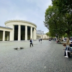 An einem bewölkten Tag gehen und sitzen die Menschen in der Nähe eines runden, weißen, neoklassizistischen Gebäudes mit hohen Säulen auf einem kopfsteingepflasterten Platz, der von grünen Bäumen gesäumt ist.