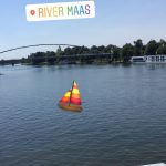 Ein ruhiger Fluss mit einer Brücke im Hintergrund, Bäumen am Ufer und einem Aufkleber mit der Aufschrift RIVER MAAS am oberen Rand. Ein buntes Segelboot-Emoji überlagert das Wasser im Vordergrund.