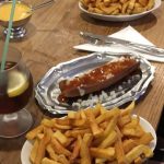 Ein Metalltablett mit einer mit Zwiebelwürfeln und Soße belegten Wurst, serviert mit einer großzügigen Portion Pommes frites auf einem Holztisch. Ein Getränk mit Zitrone und ein weiterer Teller mit Pommes frites sind ebenfalls zu sehen.