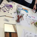 Ein Tisch mit Buntstiften, Pinseln und Papier, auf dem handgezeichnete, bunte Graffiti-Schriftzüge zu sehen sind. Ein Glas mit Pinseln und ein Behälter mit Hüttenkäse stehen inmitten des Kunstzubehörs. Die Hände der Menschen sind sichtbar.