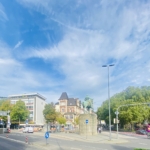 Eine städtische Straßenszene mit einer Statue einer Person auf einem Pferderücken auf einem Steinsockel. Im Hintergrund sind umliegende Gebäude, Bäume, Autos und ein teilweise bewölkter Himmel zu sehen.