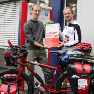 Zwei Männer stehen neben einem beladenen Tourenrad vor einem Gebäude, lächeln und halten ein Schild mit der Aufschrift Feuervogel. Das Fahrrad ist vollgepackt mit Taschen und Ausrüstung, und ein Mann trägt Sportkleidung mit einem Helm in der Nähe.