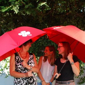 Drei Frauen, die im Freien unter zwei leuchtend roten Regenschirmen stehen, lächeln und miteinander sprechen. Üppiges grünes Efeu und Sonnenlicht bilden einen lebendigen Hintergrund.