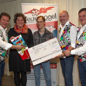 Fünf lächelnde Erwachsene stehen in einem Haus, drei Männer in bunten Westen halten einen großen Scheck. Eine Frau und ein Mann in Freizeitkleidung stehen in der Mitte. Im Hintergrund ist ein Banner mit Feuervogel zu sehen.