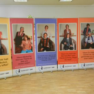Sechs bunte vertikale Banner zeigen sitzende Personen mit unterschiedlichen Gesichtsausdrücken. Auf jedem Banner steht ein deutscher Text über Alkohol, und am unteren Rand sind die Logos der EU und von Euprevent abgebildet. Die Banner stehen in einer Reihe auf einem Holzboden in einem Innenraum.