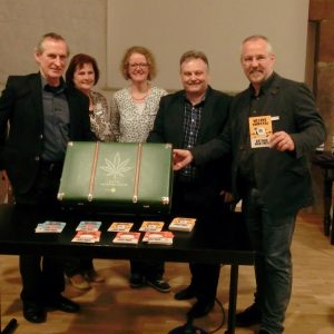 Fünf Personen stehen hinter einem Tisch, auf dem ein grüner Koffer mit einem Cannabisblattmuster und mehreren bunten Broschüren steht. Eine Person hält eine Broschüre hoch. Sie befinden sich in einem warm beleuchteten Raum mit Stein- und Holzwänden.