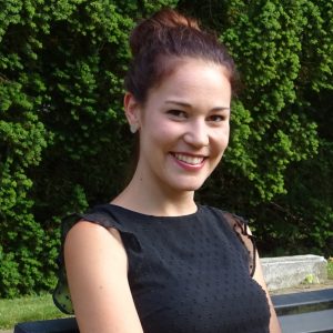 Eine junge Frau mit braunem Haar in einem Dutt und einem ärmellosen schwarzen Kleid sitzt auf einer Bank im Freien und lächelt in die Kamera. Im Hintergrund sind grüne Sträucher und Sonnenlicht zu sehen.