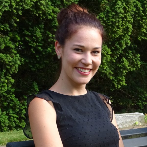 Eine junge Frau mit braunem Haar in einem Dutt und einem ärmellosen schwarzen Kleid sitzt auf einer Bank im Freien und lächelt in die Kamera. Im Hintergrund sind grüne Sträucher und Sonnenlicht zu sehen.