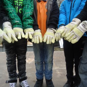 Sechs Personen, die schwere Arbeitshandschuhe tragen, stehen nebeneinander, die Arme nach vorne gestreckt und die Hände dicht beieinander. Sie sind mit legerer Outdoor-Kleidung bekleidet, darunter Jeans und Jacken.