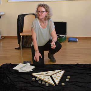 Eine Frau mit lockigem Haar kniet auf dem Boden und spricht mit Gesten. Vor ihr befindet sich eine dreieckige Tafel mit kreisförmigen Ausschnitten, Kerzen und Büchern, die alle auf einem schwarzen Tuch ausgebreitet sind. Andere Personen sind teilweise in der Nähe zu sehen.