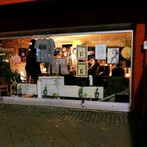 In einem gemütlichen Café mit großen Fenstern an der Vorderseite sitzen Menschen in geselliger Runde. Kunstdrucke und T-Shirts hängen im Fenster. Pflanzen und eine Flasche schmücken die Fensterbank. Draußen steht eine Person in einer hellen Jacke in der Nähe des nächtlichen Eingangs.