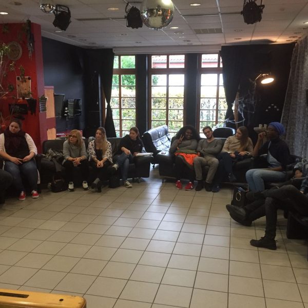 Eine Gruppe von Menschen sitzt in einem Halbkreis auf Sofas und Stühlen in einem Wohnzimmer mit gefliestem Boden, großen Fenstern und gedämpfter Beleuchtung und scheint in eine Diskussion oder Besprechung vertieft zu sein.